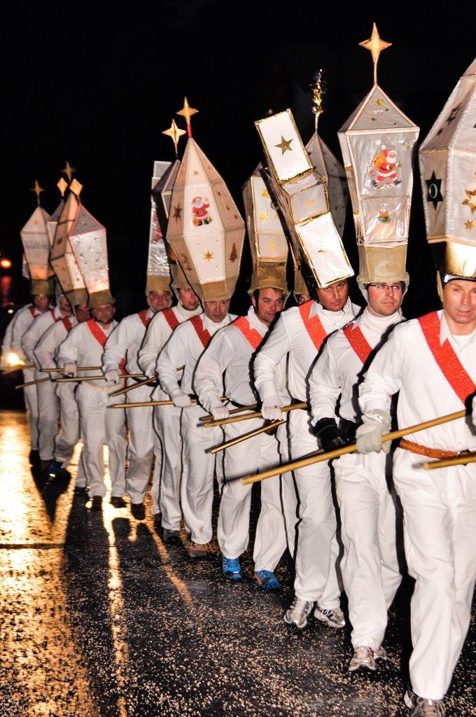 Picture of a procession of bell ringers, people wear white costumes and a large headdress made of paper
